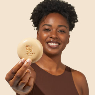Shot of model looking at the camera and smiling while holding Beekman 1802's Fresh Baked Cookies & Milk Round Bar Soap up to the camera, on a cream colored background.