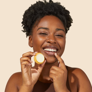 Up close shot of model holding an opened Beekman 1802's Fresh Baked Cookies & Milk lip balm and smiling, while applying the lip balm to her lips with her finger, on a cream colored background.