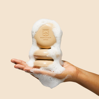 Up close image of hand holding a stack of Beekman 1802's Fresh Baked Cookies & Milk Round Bar Soap Trio, that's covered in suds on a cream colored background. 
