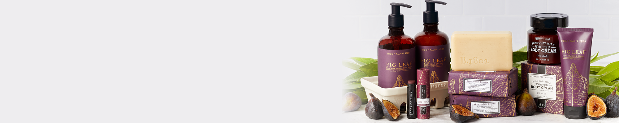 Products from Beekman 1802's Fig Leaf collection which includes the caddy set, lip balm, bar soaps, body cream, and hand cream all displayed next to each other on the far right of the screen, surrounded by sliced figs and leaves on a light grey tile background. 
