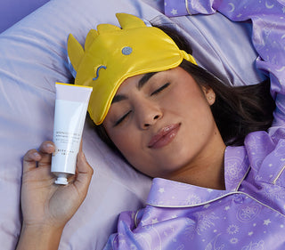 Up close shot of model dressed in purple pajamas with a yellow sleep mask on her forward, while laying on a purple pillow with her eyes closed and holding up a tube of Beekman 1802's Midnight Milk Better Aging Sleep Mask up with one hand.