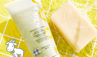 Tube of Beekman 1802's Sunshine Scrub and unwrapped bar of Beekman 1802's Goat Milk Soap laying in a pool of water with ripples, with yellow tile in the background and cartoon white goatie on the bottom left of the image.