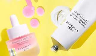 Bottle of Beekman 1802's Blotting Booster PHA & Calamine Blemish Serum and Tube of Beekman 1802's Milk Glaze Clay Mask on a yellow background, surrounded by blotting booster serum droplets and milk glaze mask product smear behind the products.