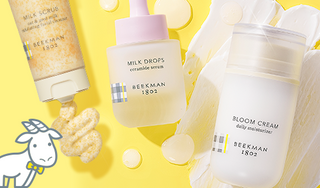 Beekman 1802's Milk Scrub Cleanser, Milk Drops Ceramide Serum, and Bloom Cream Daily moisturizer, showing squeezed cleanser out of the tube, and surrounded by serum droplets and moisturizer texture smear on a yellow background, with white cartoon goatie on the left of the screen.  