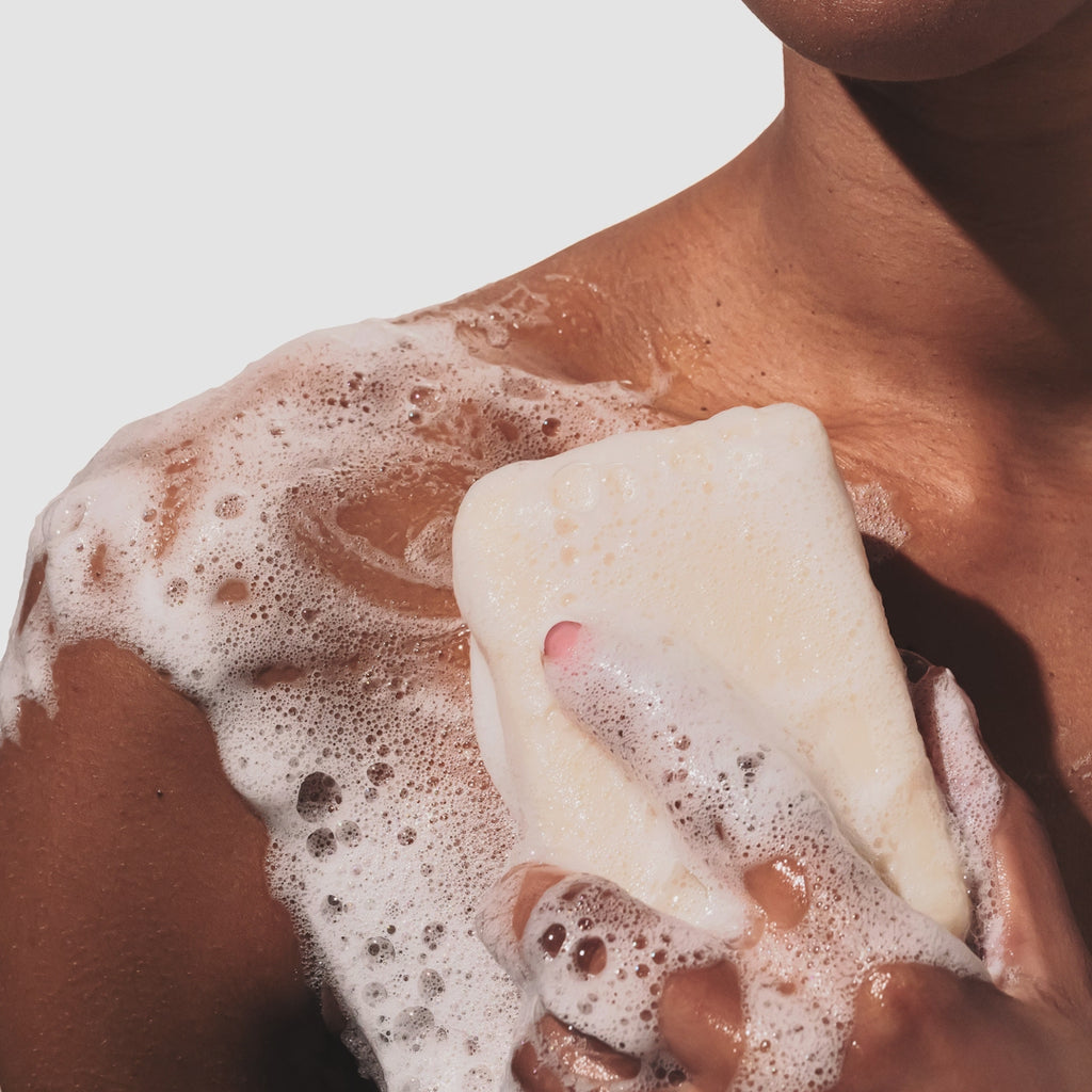 Up close GIF of model rubbing a sudsy Bar of Beekman 1802's Nestle Toll House Fresh Baked Cookies & Milk Goat Milk Soap onto their skin, creating suds, on a white background. 