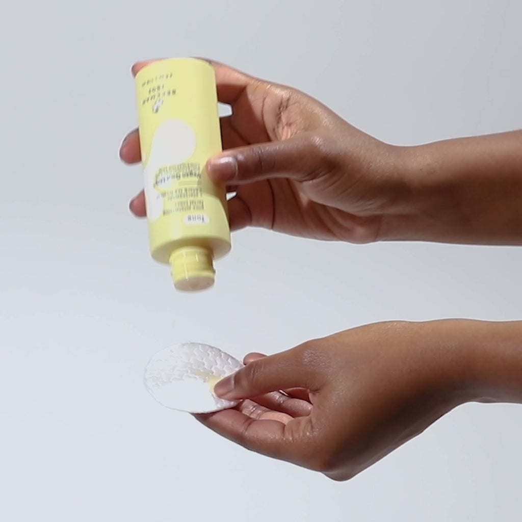 Hand holding bottle of Beekman 1802's Vegan Goat Milk™ Pore Minimizing Facial Toner and shaking the product out onto a cotton pad, on a white background. 