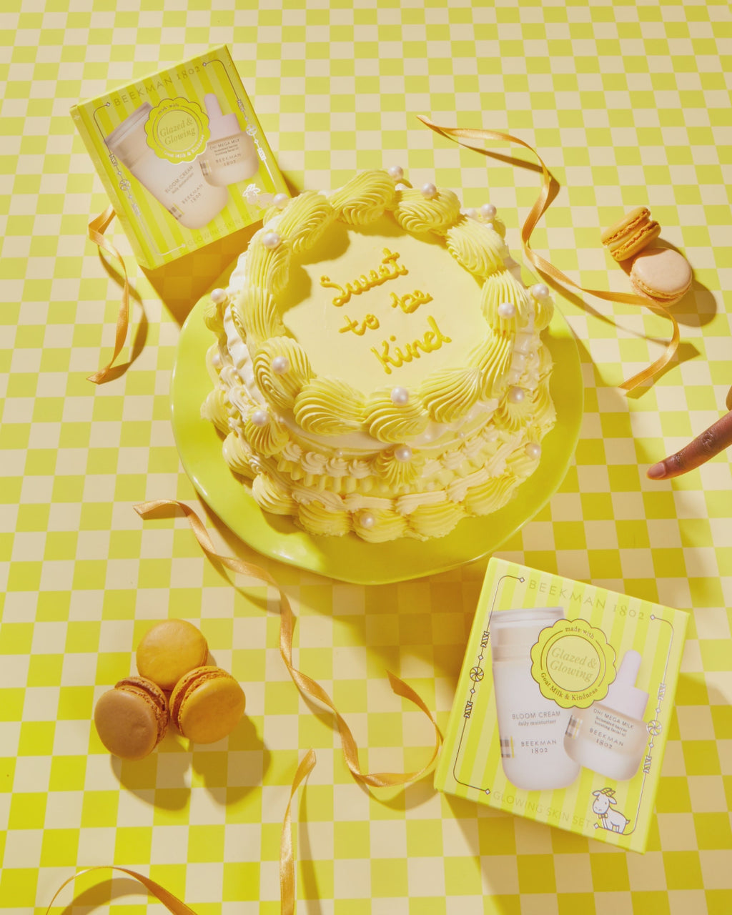 GIF of a yellow cake that says "sweet to be kind" next to two gift boxes for the Glazed & Glowing Advanced Hydration Gift set, with a hand coming from the right side and touching one of the boxes, and both of the boxes disappear to show  the Bloom cream daily Moisturizer and the Oh mega milk serum, on a yellow checkered background.