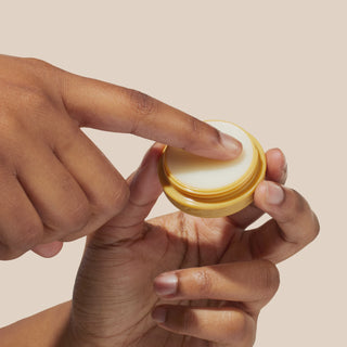GIF of model holding Beekman 1802's Beekman 1802's Fresh Baked Cookies & Milk lip balm and rubbing her finger in it, then the GIF cuts to her applying the lip balm to her lips while smiling at the camera and holding the cookie shaped lip balm container, on a cream colored background. 