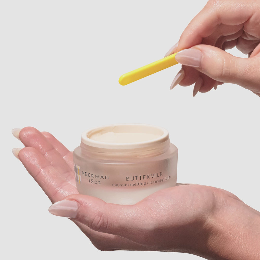 Video of a hand holding the beekman 1802 Buttermilk Makeup Melting Cleansing Balm while using the yellow spatula with the other hand to scoop the product out.