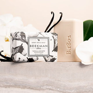 the beekman 1802 vanilla absolute wrapped bar soap next to vanilla and flowers with an unwrapped bar soap on the right.