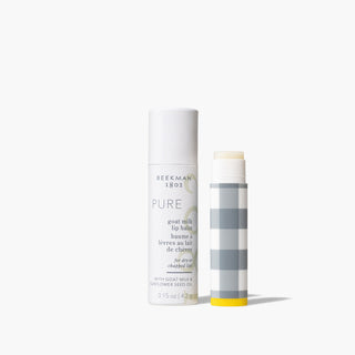 Grey and white uncapped tube with a yellow bottom of Beekman 1802's Pure Goat Milk Lip Balm standing next to a white packaging tube for the lip balm that says "Pure" on the front, on a white background. 