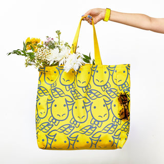 Hand holding Beekman 1802's 2023 Limited edition yellow baby goat tote with close up image of goatie's face patterned all over the bag, and tote filled with flowers with sunglasses in the pocket, on a white background. 