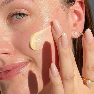 Up Close shot of side of models face while she is applying Beekman 1802's Milk Scrub Oat + Goat Milk Exfoliating Facial Cleaner to her cheek with her fingertips while looking at the camera. 