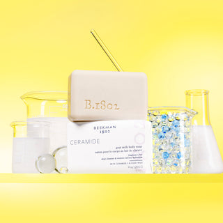 An image of an unwrapped ceramide bar soap on top of a wrapped ceramide bar soap surrounded by beakers that are filled with liquids and ingredients for the bar soap on a yellow background.