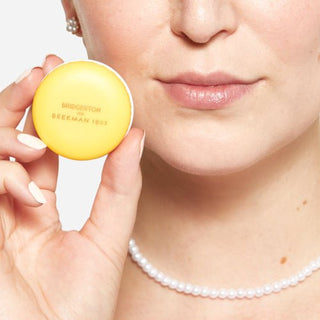 An image of a model holding up the Beekman 1802 Bridgerton Marcron Lip balm up to her face. 