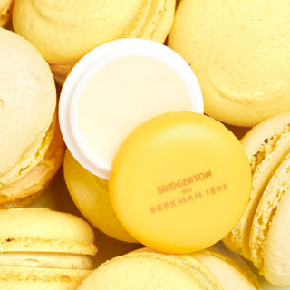 An image of the Beekman 1802 Bridgerton Macron lip balm in sunshine creme, surrounded by actual macrons. 