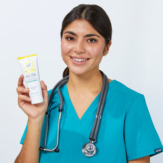 Image of a nurse in blue scrubs and with a stethoscope around her neck holding the nurses first hand cream up and smiling. 